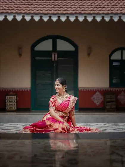 A wide shot of the bride in a stunning pink saree, seated in the courtyard of a traditional house. The reflection on the wet floor adds a beautiful symmetry to the portrait.
