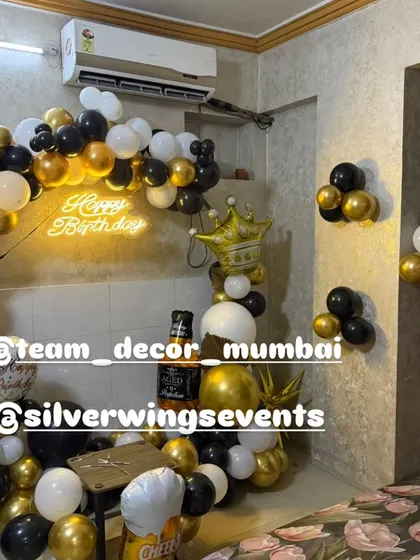 A stylish black, white, and gold themed birthday decoration for a small space. The setup includes a balloon half-arch and a 'Happy Birthday' neon sign.