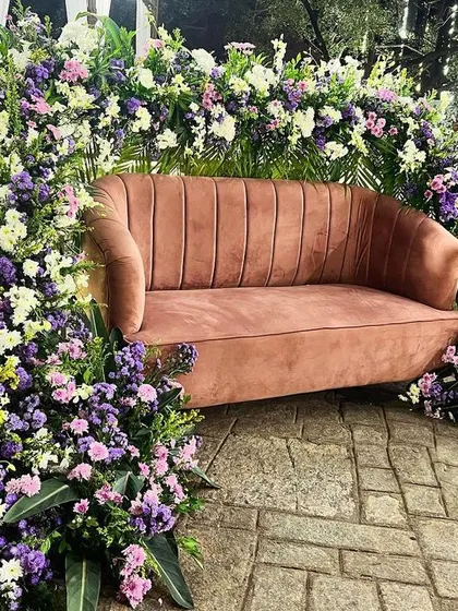 A unique photo booth for a sangeet, featuring a plush pink sofa framed by an arch of purple and white flowers.