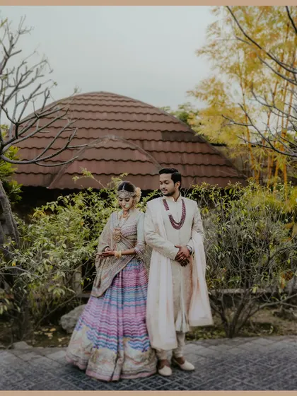 A couple stands together in a garden setting with a unique domed structure in the background. Their coordinated pastel and cream outfits look beautiful in this serene pre-wedding portrait.