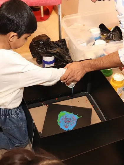 An instructor helps a young boy with a spin art project. By adding drops of paint to a spinning canvas, kids learn about centrifugal force while creating a unique masterpiece.