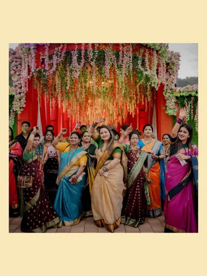 The bride's side of the family, all dressed in beautiful sarees, dance and celebrate with joy under a cascade of flowers.