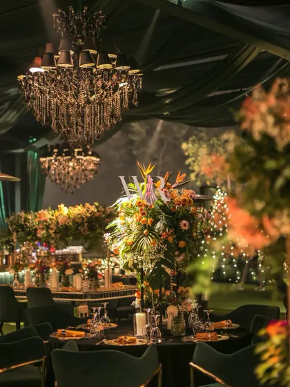 The play of light and shadow is key to creating atmosphere. Here, a spotlight catches a tall floral centerpiece, while the background glows with the soft light of chandeliers and fairy lights in the distance.