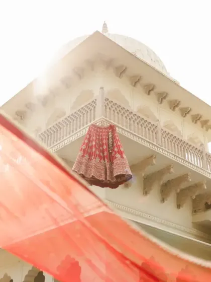 The bride's red lehenga hangs against the backdrop of a sun-kissed palace dome, with her dupatta flowing in the foreground. This artistic shot sets the stage for a royal wedding fairytale.