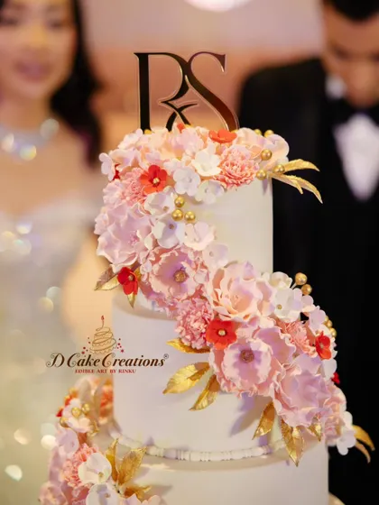 A close-up of the exquisite, handcrafted sugar flowers on a wedding cake. This shot highlights the lifelike detail of the pink peonies and red blossoms, accented with edible gold leaves and a custom monogram topper.