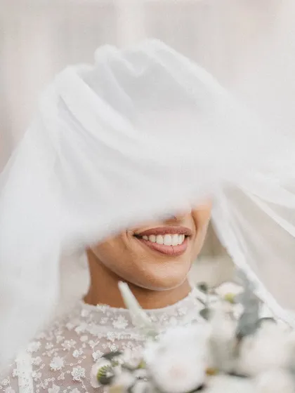 A joyful, candid close-up of a bride smiling through her veil. The movement of the veil and her bright smile create a feeling of pure, uninhibited happiness.