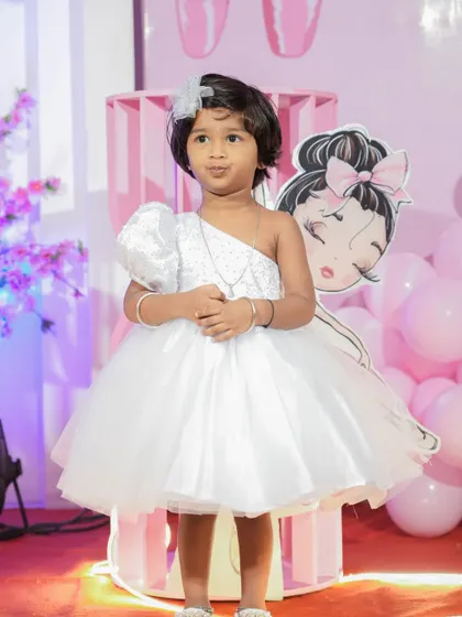 The perfect dress for a second birthday party. This custom white gown has a trendy one-shoulder design with a sparkling bodice and a multi-layered tulle skirt, ensuring the birthday girl stands out on her special day.