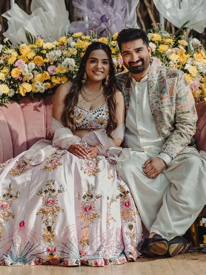 A romantic portrait of the couple at their Mehendi, seated on a pink sofa enveloped by a crescent of vibrant yellow and pink flowers.