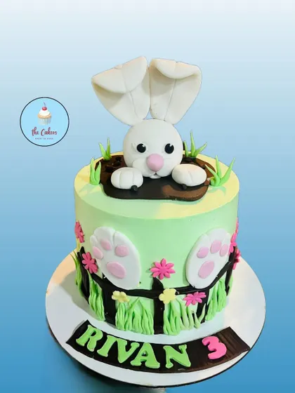 A playful bunny-themed cake for a third birthday. This cake shows a cute fondant bunny popping out of a hole, with its paws and tail visible on the sides of the cake.