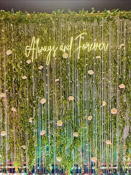 This photo backdrop brings the outdoors in with a lush wall of green foliage. I wove delicate strings of fairy lights and white roses through the leaves to create a magical, sparkling effect. The custom "Always and Forever" neon sign adds a personal and romantic touch, making it an ideal spot for capturing memories.