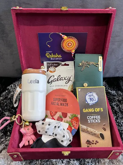 A lovely Rakhi hamper for a sister to give, featuring a personalized sipper and clutch, a face mask, coffee sticks, and other treats, all in a velvet-lined suitcase box.