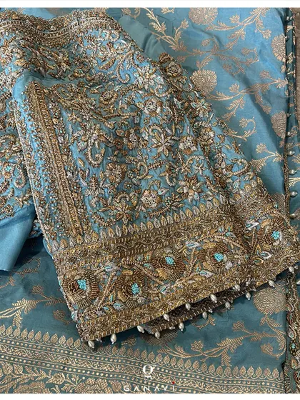An opulent teal blue blouse covered in dense zardozi and beadwork. The intricate floral patterns and pearl-drop details on the sleeves make this a true statement piece, perfect for a grand celebration.
