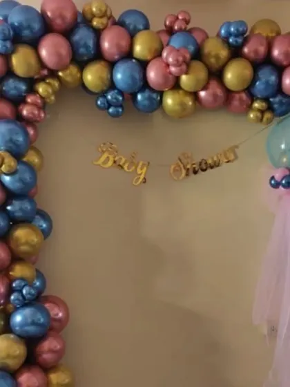 A baby shower balloon arch in a striking color combination of metallic pink, blue, and gold. The 'Baby Shower' banner hangs in the center, creating a festive frame for the celebration.