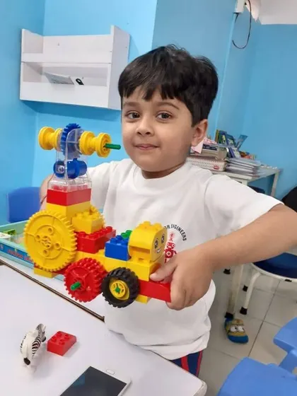 A young student shows off a vehicle he built with large, colorful bricks, a perfect introduction to mechanical concepts like gears and wheels for our youngest engineers.