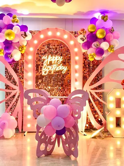 A home birthday decoration featuring a butterfly theme for an 8th birthday. The setup includes a lighted arch, large pink butterfly wings, a sequin backdrop, and a marquee number '8'.