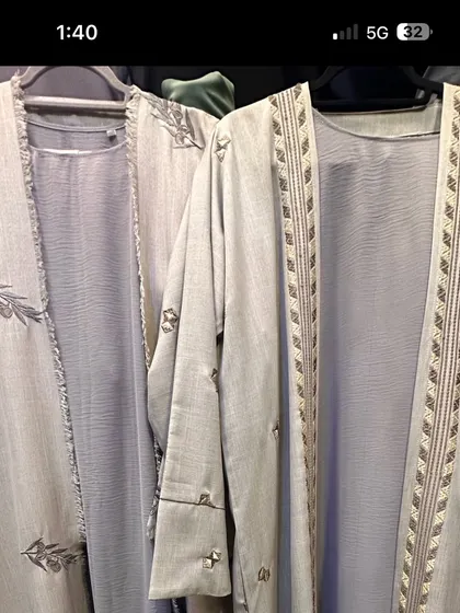 A pair of light grey open front abayas. One is detailed with a leaf pattern embroidery, while the other has a geometric design along the opening.