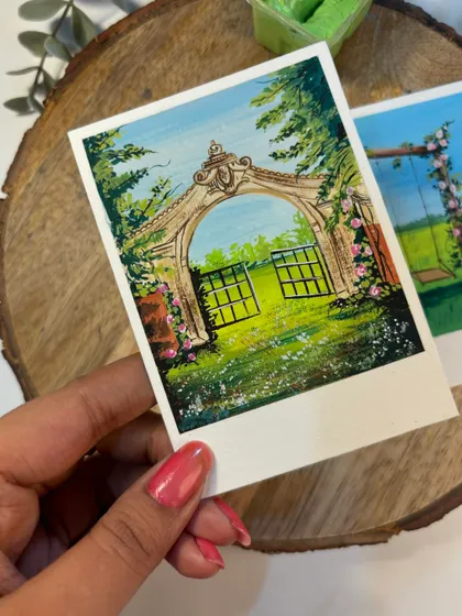 A close-up of my hand holding a polaroid painting of a whimsical garden gate. The gouache colors are so vibrant against the green background.