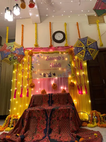 A cozy at-home Haldi setup for 'Zainab's Haldi'. The decor uses yellow drapes, marigold garlands, and Rajasthani umbrellas to transform a living room into a festive space.