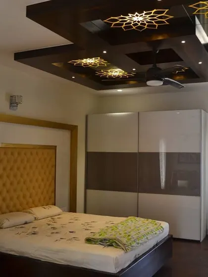 This bedroom combines classic and modern elements, with a tufted yellow headboard set against a white high-gloss panel. The highlight is the intricate false ceiling, which features dark wood panels with backlit, laser-cut floral motifs.
