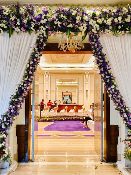 An elegant floral archway at the entrance of a grand ballroom. The combination of purple and white flowers with draped white fabric creates a sophisticated and welcoming feel.