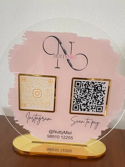A stylish, circular acrylic stand for 'NuttyMiel' featuring a pink brushstroke design, their logo, and gold-framed QR codes for Instagram and payment.