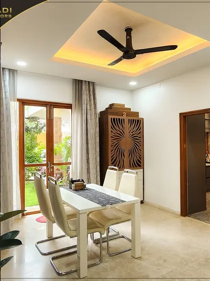 A bright dining area situated by a large window overlooking a garden. The simple white dining table and chairs are complemented by a custom wooden cabinet with intricate jali work.