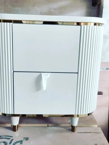 A custom-made bedside table in our workshop. The design features fluted side panels, a white deco paint finish, and elegant gold-plated legs and top trim.