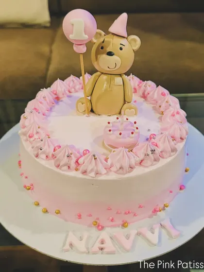 A sweet and simple first birthday cake in pink. It's topped with an adorable fondant teddy bear holding a "1" balloon, with the name "Navi" written in pink letters.
