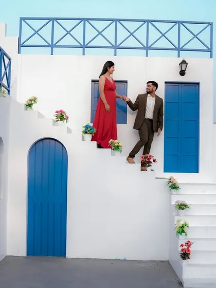 A fairytale moment on the steps of a beautiful Santorini-style building. The groom helps his bride-to-be, dressed in a flowing red gown, creating a classic and romantic cinematic shot.
