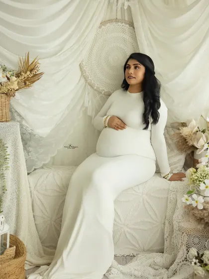 A serene and thoughtful portrait of a mother-to-be seated on a daybed. The all-white setting creates a peaceful and heavenly atmosphere.