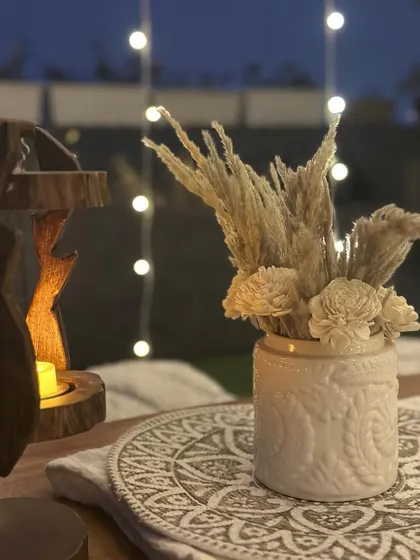 Cozy and boho vibes for a birthday soiree. This close-up shows the details of the tablescape, including dried pampas grass in a textured vase and a wooden incense holder, all against a backdrop of fairy lights.