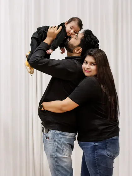 A loving family of three captured in a candid moment, with the father lifting their baby high as the mother hugs him from behind, all sharing a beautiful connection.