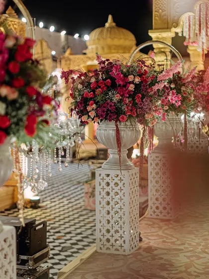 The grand aisle leading to the mandap, lined with large floral urns on illuminated pedestals. The checkered floor and the distant stage create a sense of anticipation and grandeur.