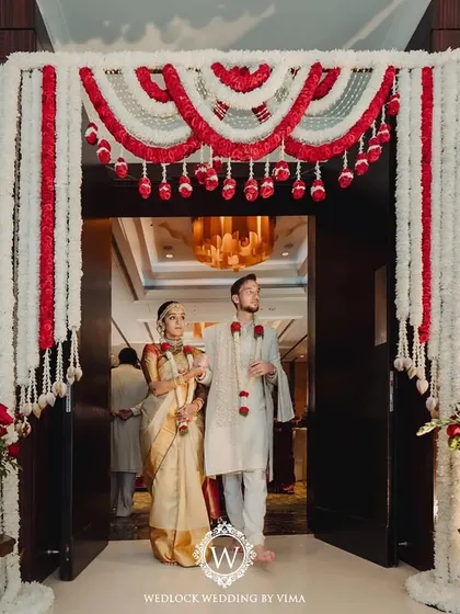 The couple's entrance through a grand doorway framed by traditional red and white floral garlands. This setup created a beautiful and dramatic start to their wedding ceremony.