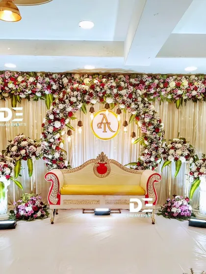 This engagement stage features a beautiful floral arch made of red, white, and purple blooms, with a custom initial monogram hanging in the center. The warm drop lighting adds a soft, romantic glow to the setting.