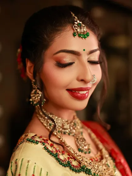 A close-up portrait of a model with a gentle, happy smile. Her eyes are closed, drawing attention to her shimmering eyeshadow and the intricate details of her bridal jewelry.