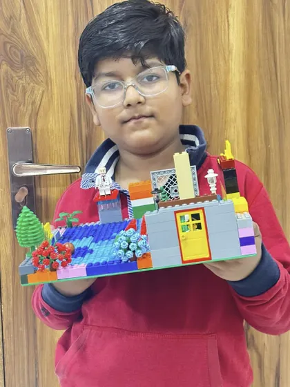 A student proudly holds up his completed LEGO house, a project that teaches basic architectural concepts and design.