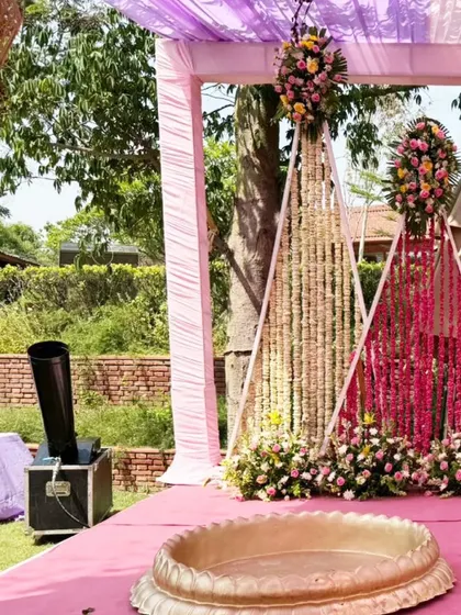 The Haldi ceremony stage, set against a backdrop of hanging floral strings in shades of pink and white. The traditional brass 'urlis' are ready for the ritual.