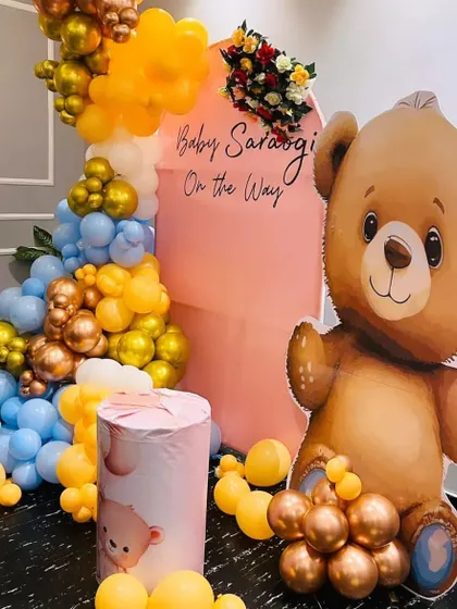 A cute teddy bear themed baby shower with a "Baby On The Way" message. This setup includes a large teddy bear cutout, a pink arched backdrop, and a colorful balloon arrangement in blue, yellow, and gold.