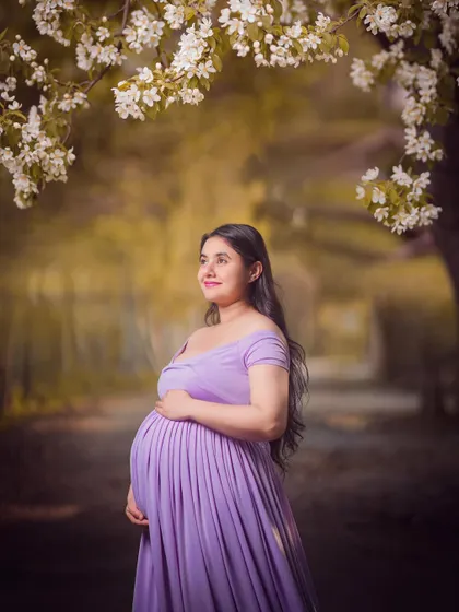 A serene portrait of a mom-to-be in a flowing lavender dress, standing under a canopy of white blossoms. The soft light and dreamy background create a peaceful and beautiful scene.