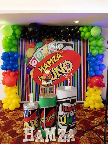 A unique UNO-themed first birthday party. The backdrop and plinths are designed with the iconic card game's colors and symbols, framed by a balloon arch in red, yellow, green, and blue.