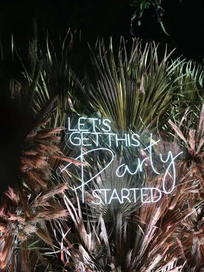 "Let's Get This Party Started." A fun neon sign set against palm trees, instantly setting a celebratory mood for the Sangeet night in Alibaug.