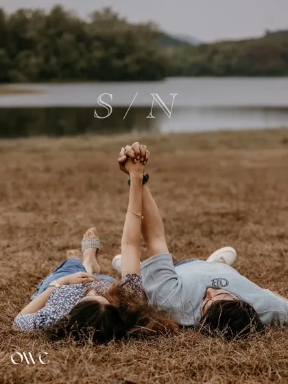 A relaxed pre-wedding moment with the couple lying on the grass, holding hands against a scenic lake backdrop.