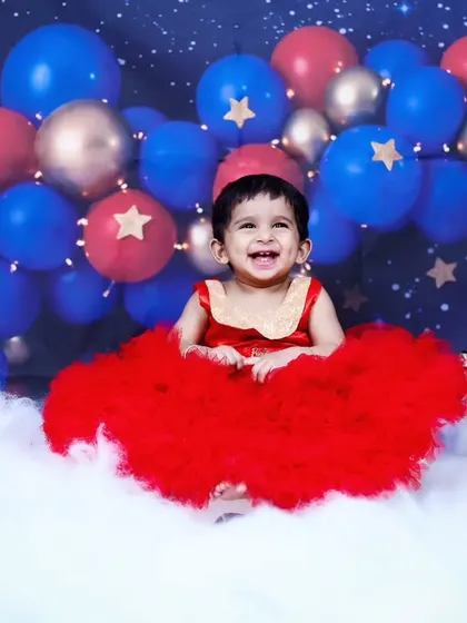 A magical fairy-themed shoot with a baby girl in a stunning red dress, sitting on a cloud under a canopy of balloons and stars.