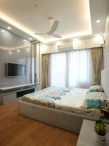 A full view of the bedroom, showcasing the elegant grey and gold accent wall, the cozy bed, and a stylish TV unit with a vertical-striped panel backdrop.