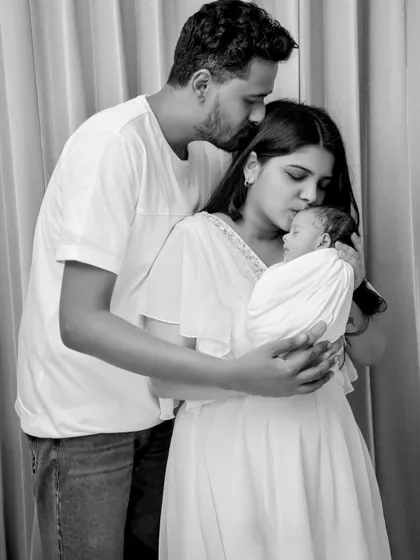 A beautiful black and white image of a family of three. The father kisses his wife's forehead as she cradles their newborn, capturing a serene and loving moment.