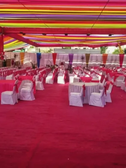 A large-scale pandal decoration with a ceiling of colorful rainbow drapes. The entire floor is carpeted in red, and the space is filled with tables and chairs for guest dining.