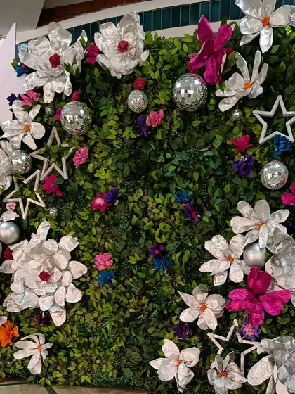 A unique and artistic photo wall for a modern reception. The wall of green foliage is decorated with oversized paper flowers, stars, and disco balls, creating a fun and glamorous backdrop.