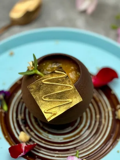 A sphere of caramelised chocolate mousse sits in a pool of rich chocolate sauce, topped with a delicate gold leaf square. It's a dessert that exudes luxury and sophistication.