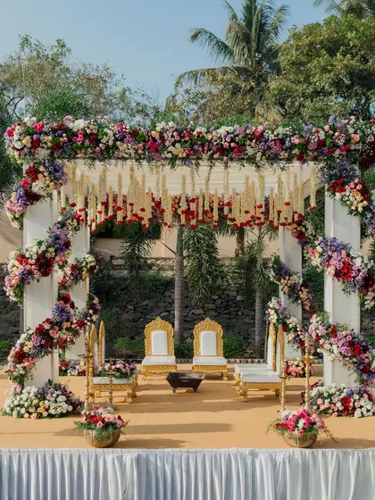 The vibrant, multi-colored floral mandap stands ready for the ceremony, with traditional golden chairs awaiting the bride and groom.